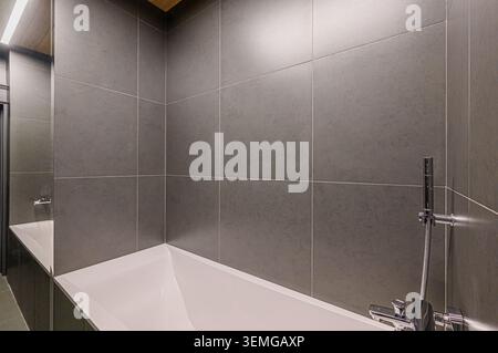 Moderne Badezimmerecke mit weißer Badewanne und grauen Fliesenwänden mit Chromarmaturen. Klares und minimalistisches Design Stockfoto
