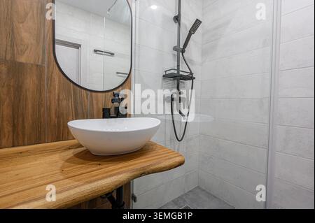 Modernes Bad mit weißem Waschbecken auf einer Holzarbeitsfläche, mit schwarzem Wasserhahn und Spiegel. Dusche mit weißen Fliesen und schwarzen Armaturen Stockfoto
