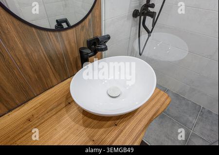 Der moderne Kosmetikbereich im Bad verfügt über ein weißes Keramikwaschbecken, einen schwarzen Wasserhahn und eine Holzarbeitsfläche. Die Wand hinter dem Waschbecken ist gefliest Stockfoto