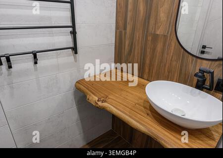 Stilvolle Badezimmerecke mit Holzarbeitsfläche mit weißem ovalen Waschbecken, schwarzen Armaturen und gefliesten Wänden in hellgrauen und holzähnlichen Texturen Stockfoto