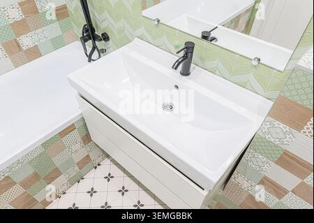 Das helle Badezimmer bietet Blick auf ein weißes Waschbecken mit schwarzem Wasserhahn, gemusterte grüne und holzfarbene Fliesen und einen mit Blumenfliesen verzierten Boden. Ein mirr Stockfoto