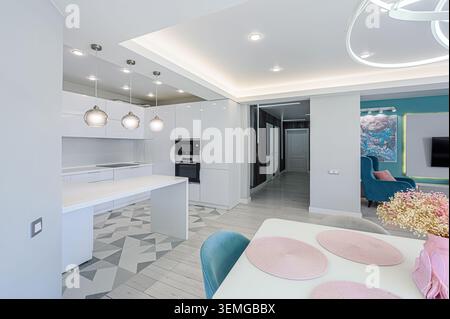 Das helle, moderne Apartment verfügt über eine elegante, weiße Küche, einen Essbereich mit pinkfarbenen Akzenten und einen Wohnbereich mit blauer Wand und blauem Armchai Stockfoto