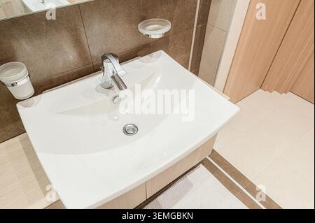Sauberes, gut beleuchtetes Waschbecken im Bad mit einem Chromhahn und einer Seifenschale an der gefliesten Wand. Das Waschbecken ist weiß und die Umgebung ist neutral Stockfoto