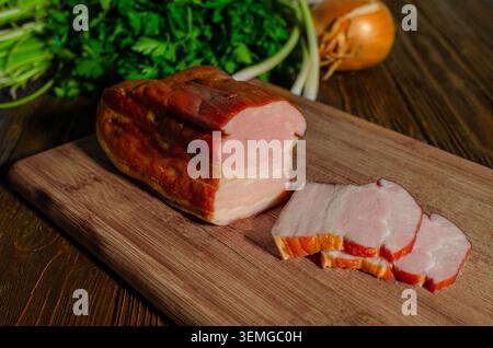 Ein rustikales Stück geräuchertes Schweinebauchbrust neben frischen Scheiben auf einem Holzschneidebrett, mit frischem Petersilie und Zwiebeln auf dem Rücken Stockfoto