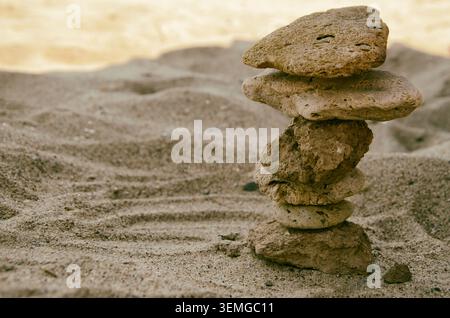 Eine Nahaufnahme eines kleinen, sorgfältig ausbalancierten Stapels rauer, poröser Meeressteine, die eine Zen-Pyramide auf dem strukturierten Sand eines sonnigen Strandes bilden, Symbol Stockfoto