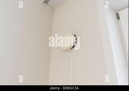 Weißer, runder Rauchmelder ist an einer weißen Wand in einem Raum montiert. Kabel hängen am Gerät. Ein Teil einer Tür ist zu sehen. Stockfoto