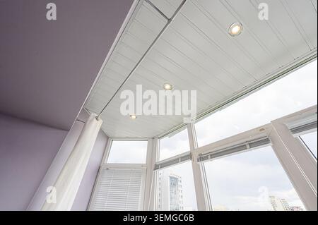 Flache Aufnahme einer Ecke eines Zimmers mit weißer Decke, Einbaubeleuchtung, großen Fenstern mit Jalousien und einer hellvioletten Wand. Stockfoto