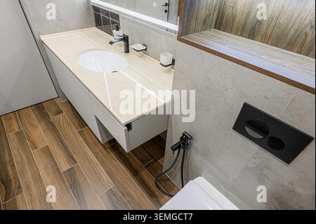 Modernes Bad mit beigefarbener Ablagefläche mit ovalem Waschbecken, schwarzem Wasserhahn und Fliesenboden in Holzoptik. Graue Fliesenwände runden das elegante Design ab Stockfoto