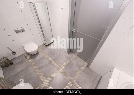 Hochwinkelaufnahme eines modernen Badezimmers mit weißen Fliesen, grauen Bodenfliesen mit dekorativem Gittermuster, Toilette, Spiegel und grauer Tür Stockfoto
