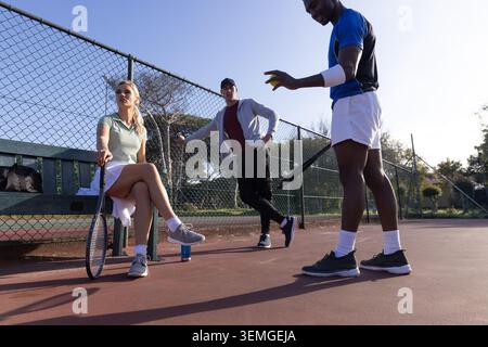 Verschiedene Freunde in Tenniskleidung sitzen auf der Hofbank, lehnen sich am Zaun, halten Schläger und Ball Stockfoto