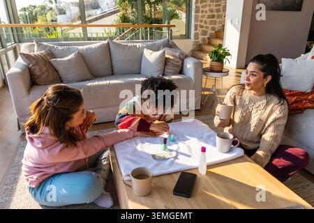Zwei Erwachsene Frauen mit afroamerikanischem Kind sitzen im Wohnzimmer am Couchtisch und malen Nägel Stockfoto