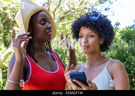 Afroamerikanische Frauen, die im Park sprechen, haben eine Frau mit Strohhut als Freundin mit blauem Smartphone verlassen Stockfoto