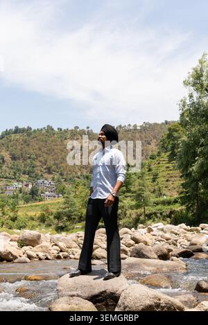 Ein vertikaler Blick auf die gesamte Länge eines jungen Sikh-Mannes in formeller Kleidung, der selbstbewusst auf Felsen über einem Bergbach in Himachal Pradesh, Indien, steht. Stockfoto