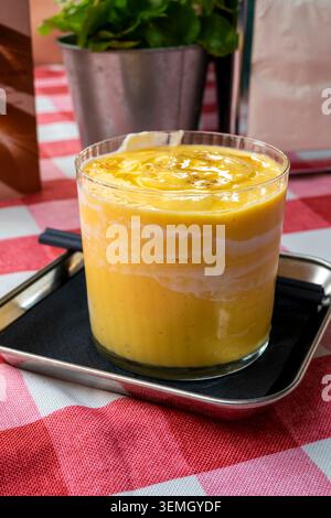 Ein cremiger Mango-Lassi mit wunderschönen Joghurtwirbeln serviert in einem Glas auf einer rot-weiß karierten Tischdecke. Stockfoto