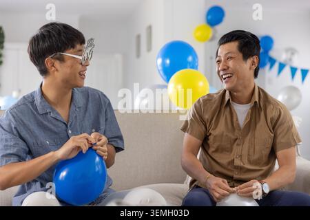 Zwei asiatische Männer sitzen auf einem hellen Sofa im Wohnzimmer und binden Ballons, einer trug eine neuartige Brille Stockfoto
