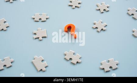 Abgewinkelte Ansicht eines orangefarbenen Fragezeichensymbols, umgeben von losen weißen Puzzleteilen auf einer sauberen blauen Oberfläche Stockfoto