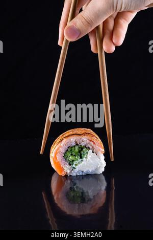 Hölzerne Essstäbchen heben eine Sushi-Rolle mit frischem Lachs aus philadelphia auf glänzend schwarzer Oberfläche Stockfoto