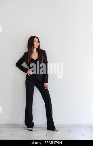 Frau, die Seite in schwarzem formellem Hosenanzug oder Anzug sieht, steht im Studio vor der weißen Wand. Kaukasisches Brünette-Modell mit langen Haaren. Unternehmensporträt Stockfoto