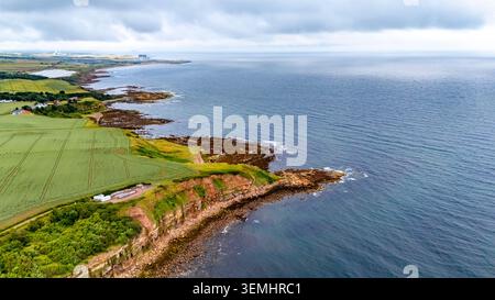 Blick aus der Vogelperspektive auf die zerklüftete Küste in Cove, Schottland, mit Blick auf die dramatischen Klippen, die felsige Küste und die üppigen grünen Felder, die sich bis zu den s erstrecken Stockfoto