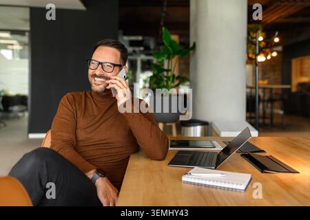 Ein lächelnder Mann mit Brille und Rollkragenhemd, der sich in einem glücklichen Telefongespräch einlässt, während er an einem Laptop in einem Co-Team arbeitet und arbeitet Stockfoto