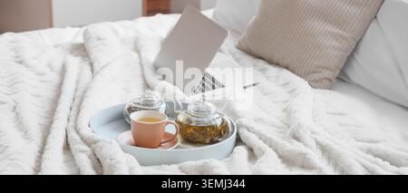 Gemütliche Schlafzimmerszene mit Laptop und Kräutertee auf Tablett über weicher weißer Decke, komfortabler Arbeitsplatz für Fernarbeit, Entspannung, Selbstpflege, Hygg Stockfoto