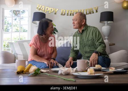 Asiatisches Paar mit einer älteren Frau, die auf dem Sofa unter dem HAPPY BIRTHDAY Banner sitzt, Kuchen und Tassen teilt Stockfoto