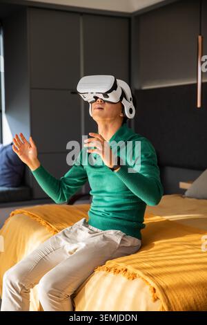 Asiatischer Mann mit grünem Pullover, weißem VR-Headset, Gesten im Schlafzimmer auf dem Bett mit Senfüberwurf Stockfoto