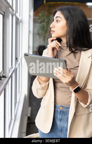 Erwachsene indische Frau, die am Fenster im Büro steht, Blazer trägt und Tablette mit Koffer hält Stockfoto