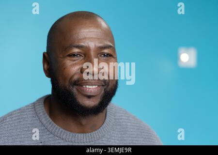 Afroamerikaner lächelt, während er mit grauem Pullover und Wandleuchte vor einer türkisfarbenen Wand posiert Stockfoto
