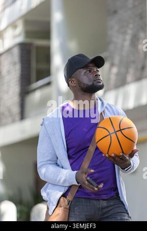 Erwachsener afrikanischer Mann, der beim Bauen steht, Mütze und Hoodie trägt, Basketball und Smartphone hält. Stadt, Straße, Freizeit, Person, Gerät, Balkon, bri Stockfoto