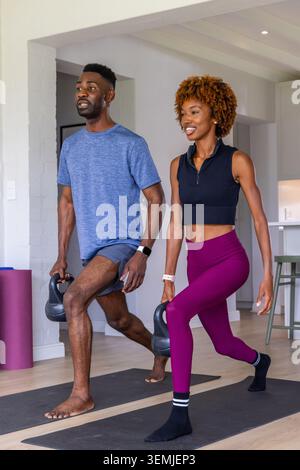 Afroamerikanisches Paar, das zu Hause eine Kettlebell-Ausfallschritte in Trainingsausrüstung auf schwarzen Matten macht Stockfoto