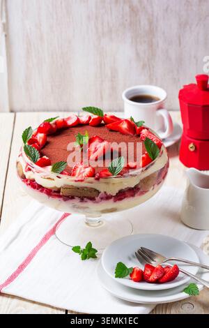 Mehrschichtiges Dessert mit Erdbeeren, Sahne und Biskuitkuchen, serviert in einer Glasschale, belegt mit frischen Beeren und Minze, gestylt auf einem hellen Holztisch wi Stockfoto