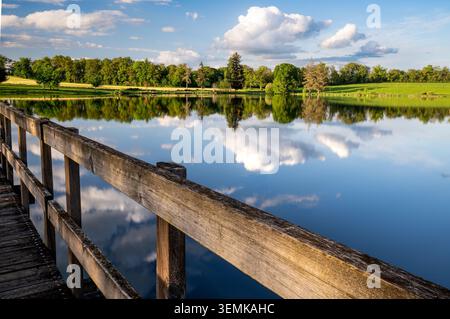 Das hölzerne Geländer eines Piers führt im Vordergrund den Blick über das ruhige Wasser des Sees Les Pouyades. Die Oberfläche spiegelt das große weiß perfekt wider Stockfoto