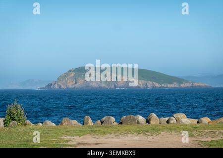 Malerischer Blick auf den Leuchtturm von Illa Coelleira auf einer grünen Insel, über das blaue Meer vom Hafen von Bares in Galicien, Spanien, unter klarem Himmel. Stockfoto