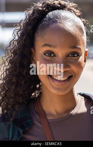 Afroamerikanerin lächelt und sieht die Kamera draußen an und trägt ein grünes kariertes Hemd, braunes Oberteil-Taschenband Stockfoto