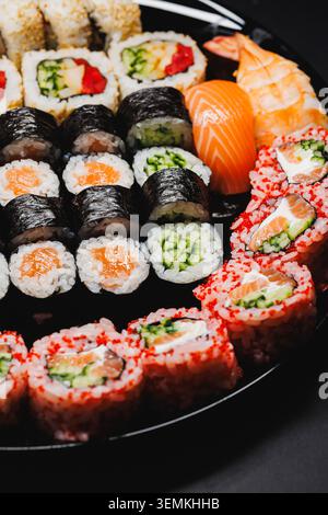Verschiedene Sushi-Brötchen sind auf einer schwarzen runden Platte angeordnet. Verschiedene Arten von Sushi sind Thunfisch, Lachs und Gurkenrollen. Leuchtende Farben ziehen Aufmerksamkeit auf sich Stockfoto