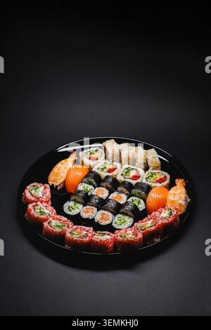 Verschiedene Arten von Sushi werden ordentlich auf eine schwarze Platte gelegt. Die Platte enthält farbige Brötchen, Lachsstücke und Wasabi. Diese Einstellung ist in Innenräumen Stockfoto