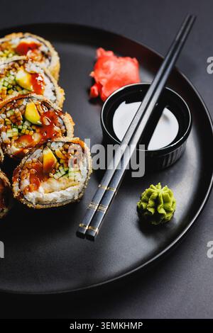 Sushi-Brötchen werden ordentlich auf eine schwarze Platte mit Stäbchen daneben gelegt. Es gibt kleine Schüsseln mit Soße, Wasabi und Ingwer, die um die Rolle angeordnet sind Stockfoto