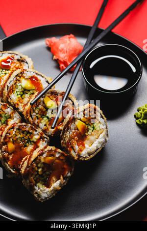 Sushi-Brötchen sind ordentlich auf einem schwarzen Teller angeordnet. Eine kleine Schüssel Sojasauce steht neben den Brötchen. Wasabi und Ingwer sind in der Nähe. Stäbchen AR Stockfoto