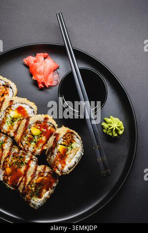 Sushi-Brötchen sind ordentlich auf einer runden schwarzen Platte angeordnet. Neben den Brötchen befinden sich rosa Ingwerscheiben, grüner Wasabi und eine kleine Schüssel Sojasauce. Zwei Cho Stockfoto