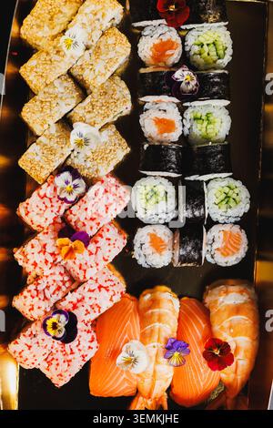 Eine Platte fasst verschiedene Arten von Sushi-Brötchen, die ordentlich angeordnet sind. Einige Brötchen haben eine Schicht Fisch, andere enthalten Gemüse. Blumen fügen Farbe hinzu Stockfoto
