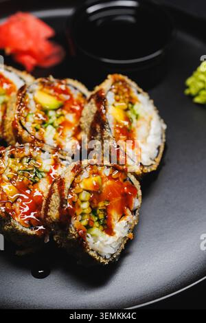 Sushi-Brötchen sind ordentlich auf einer runden schwarzen Platte angeordnet. Die Brötchen werden mit frischen Zutaten gefüllt und mit Soße belegt. Eine kleine Schüssel Sojasauce, Stockfoto