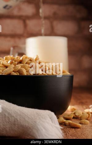 Gebackene Erdnüsse in einer schwarzen Schüssel mit einer weißen Serviette im Vordergrund und Bier wird in einem Milchbecher mit einer Backsteinmauer im Hintergrund serviert. Verti Stockfoto