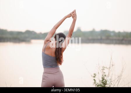 Rückansicht einer jungen Frau, die bei Sonnenaufgang Yoga im Freien macht, mit erhobenen Armen an einem ruhigen Seeufer, die Wellness, Freiheit, Achtsamkeit, Re zum Ausdruck bringt Stockfoto