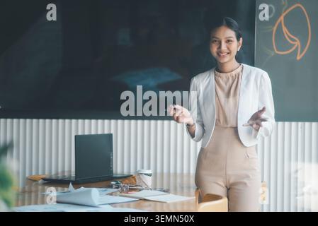 Selbstbewusste Geschäftsfrau, die neben Laptop und Dokumenten im Besprechungsraum im Büro steht und lächelt, während sie Ideen vorstellt, Diskussionen leitet und erklärt Stockfoto