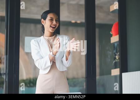 Lachende Geschäftsfrau steht und applaudiert im Büro, feiert Projekterfolg, Teamleistung und positives Feedback nach einer Präsentation in Stockfoto