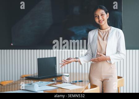 Lächelnde Geschäftsfrau präsentiert sich neben dem Bürotisch, mit geöffneten Gesten, um Ideen zu erklären, Diskussionen zu führen und die Geschäftsstrategie in A einzuführen Stockfoto