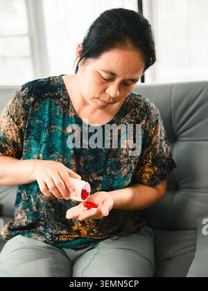 Asiatische Frau, die rote Pillen aus einer Flasche in ihre Hand gießt, während sie auf einem Sofa sitzt, repräsentiert Medikamentenroutine, Gesundheit, Behandlung, Wellness, h Stockfoto