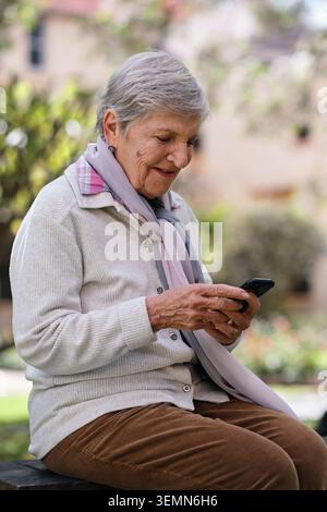 Lächeln, Bank und alte Frau im Park mit Telefon, sozialen Medien und Website für Wochenendausflüge. Glückliche, ältere Person und SMS in der Natur mit Stockfoto