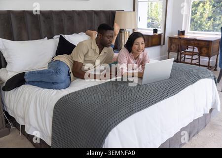 Asiatische Frau und Afroamerikaner liegen im Bett, tragen Hemden, benutzen silbernen Laptop, halten Karte Stockfoto
